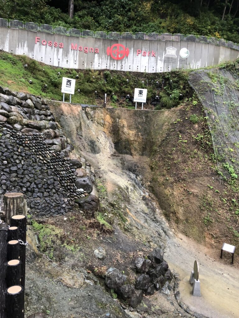 Bird's-eye view of Fossa Magna Park's fault outcrop from the upper viewing platform, showing the full scale of the excavated geological boundary between Eastern and Western Japan, with the Fossa Magna Park UNESCO Geopark sign visible on the retaining wall.