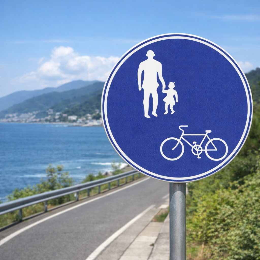 A blue circular road sign in Japan indicating a shared pedestrian and bicycle path, with icons of an adult and child walking and a bicycle, set along a coastal road with the ocean and town in the background.