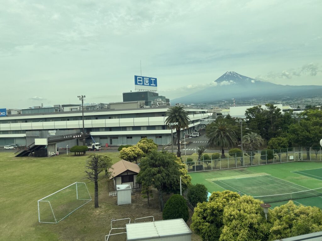 Mount Fuji rising above a layer of clouds as seen from a Shinkansen train, with green fields and rural scenery in the foreground.