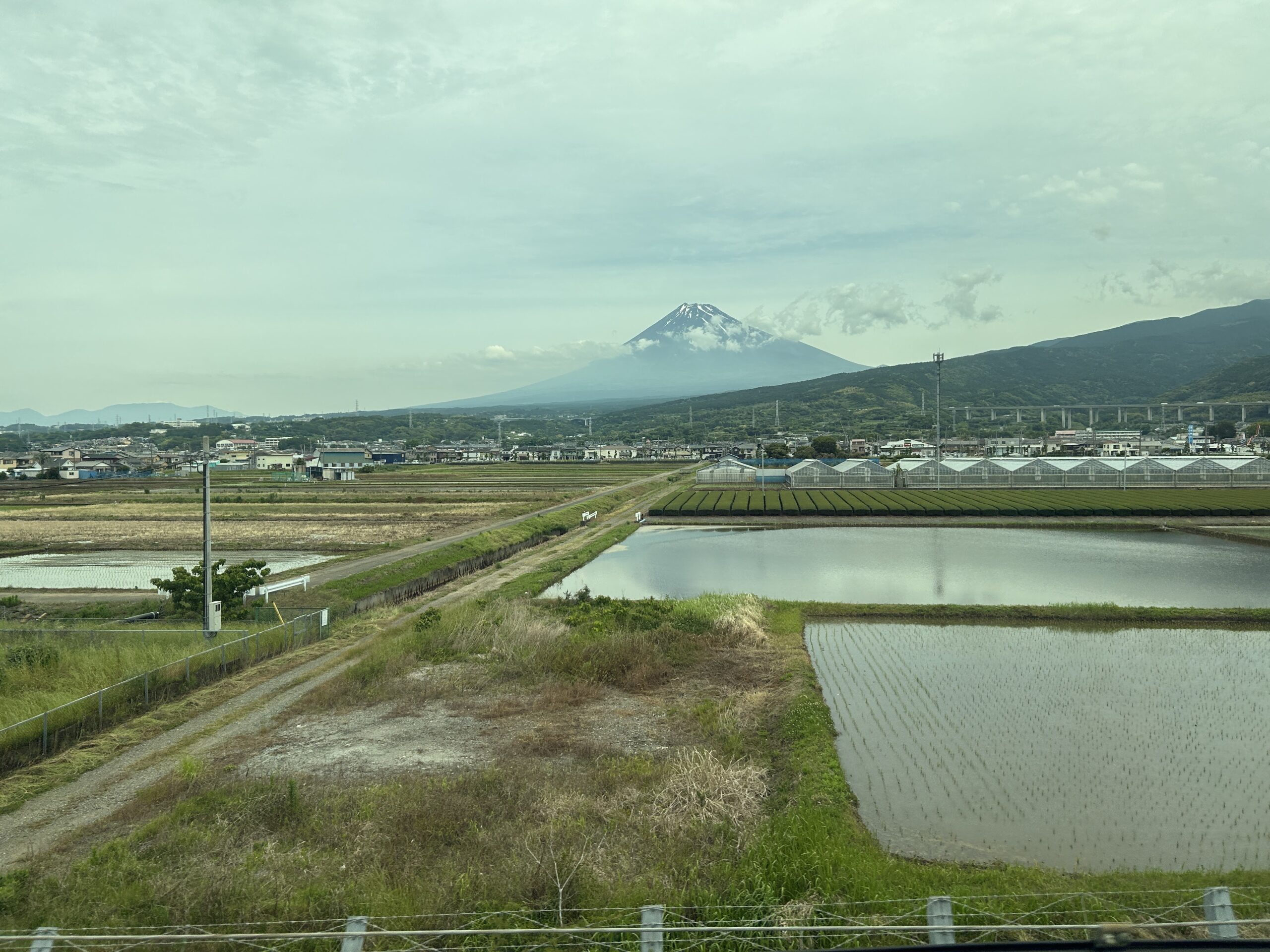 Mount Fuji above a blanket of clouds with rice fields and Japanese countryside seen from a Shinkansen E seat.